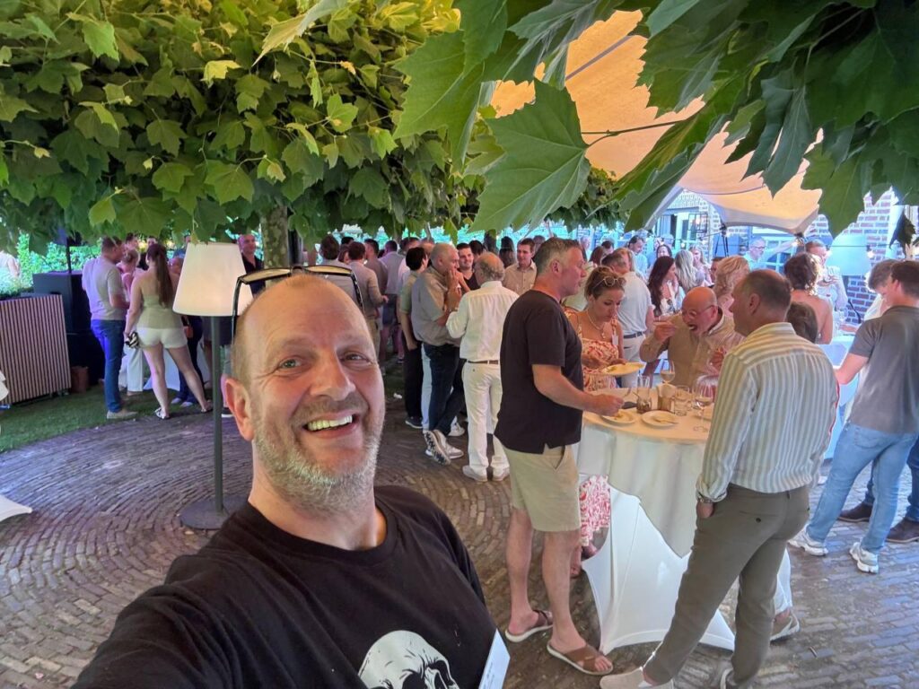 Richard maakt een selfie op de Vick Haringparty, onder de platanen, met op de achtergrond ondernemers aan statafels met hapjes en wijn.