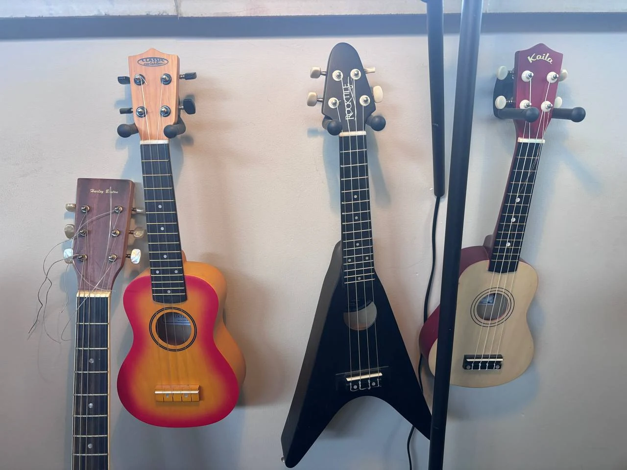 Vier kleine snaarinstrumenten aan een muur: een Harley Benton met loshangende snaren, een rood-oranje Classic Collection ukulele, een matzwarte Rocktile in Flying-V-vorm en een naturelkleurige Kaila